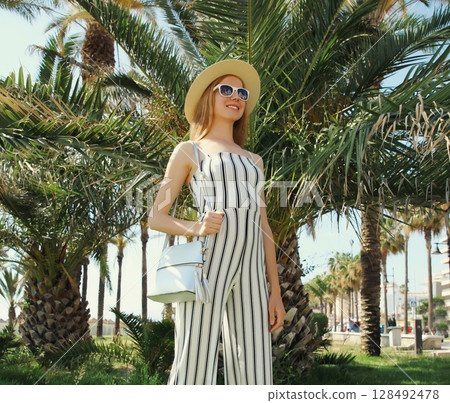 Summer outfit, stylish beautiful young woman posing in white striped jumpsuit in park, palm trees Summer outfit, stylish beautiful young woman posing in white striped jumpsuit in park, palm trees 128492478