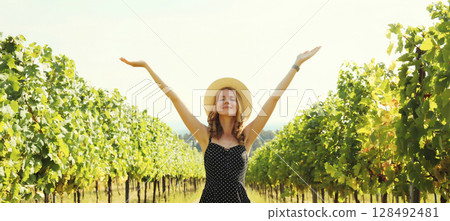 Happy young woman tourist in a summer hat enjoying sunny view of a vineyard in Italy 128492481