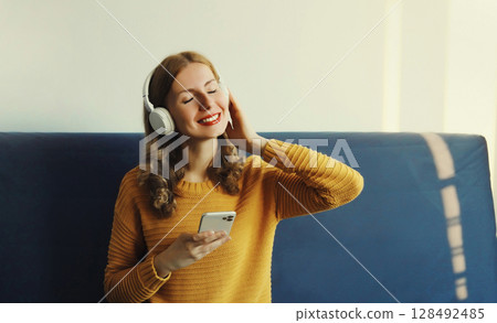 Happy smiling young woman listening to music in headphones with phone sitting on a couch at home 128492485