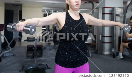 Ordinary woman in the gym doing exercises with dumbbells. Workout for weight loss. Body strengthening and healthy lifestyle 128492520