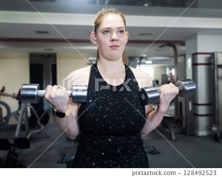 Ordinary woman in the gym doing exercises with dumbbells. Workout for weight loss. Body strengthening and healthy lifestyle Ordinary woman in the gym doing exercises with dumbbells. Workout for weight loss. Body strengthening and healthy lifestyle 128492523