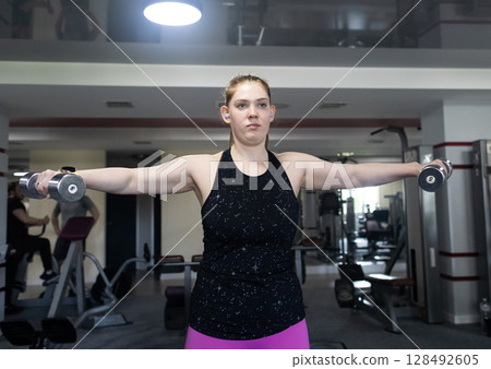 Ordinary woman in the gym doing exercises with dumbbells. Workout for weight loss. Body strengthening and healthy lifestyle Ordinary woman in the gym doing exercises with dumbbells. Workout for weight loss. Body strengthening and healthy lifestyle 128492605