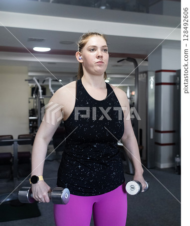 Ordinary woman in the gym doing exercises with dumbbells. Workout for weight loss. Body strengthening and healthy lifestyle Ordinary woman in the gym doing exercises with dumbbells. Workout for weight loss. Body strengthening and healthy lifestyle 128492606