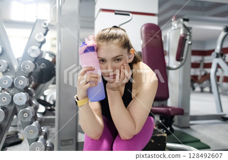 Feeling unwell in the gym. Woman tired from exercising in the gym and drinking water from a bottle and resting. Workout for weight loss. 128492607