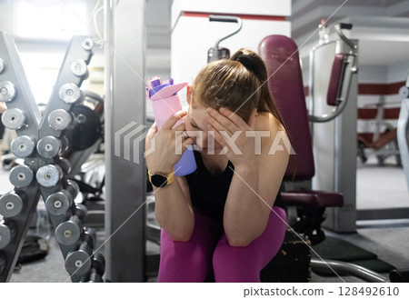 Feeling unwell in the gym. Woman tired from exercising in the gym and drinking water from a bottle and resting. Workout for weight loss. 128492610