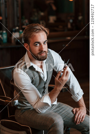 Holding scissors. Handsome barber is in the salon Holding scissors. Handsome barber is in the salon 128492717