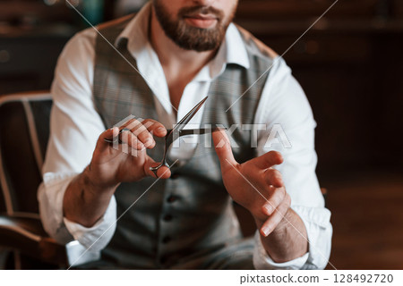 Holding scissors. Handsome barber is in the salon 128492720