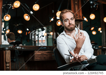 Standing and holding scissors. Handsome barber is in the salon Standing and holding scissors. Handsome barber is in the salon 128492745