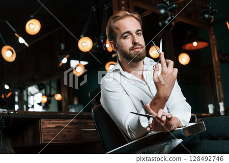 Standing and holding scissors. Handsome barber is in the salon Standing and holding scissors. Handsome barber is in the salon 128492746