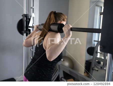 Plus size woman doing exercises on Smith Machine. Squats on a simulator with a barbell. Workout for weight loss 128492894