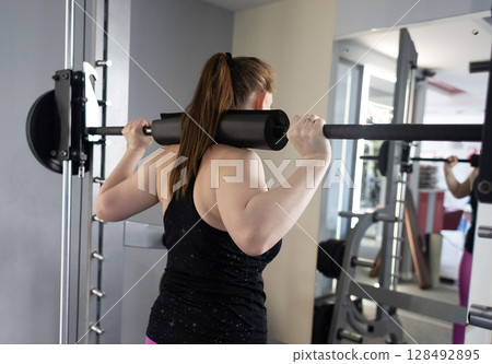 Plus size woman doing exercises on Smith Machine. Squats on a simulator with a barbell. Workout for weight loss 128492895