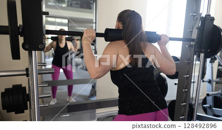 Plus size woman doing exercises on Smith Machine. Squats on a simulator with a barbell. Workout for weight loss 128492896