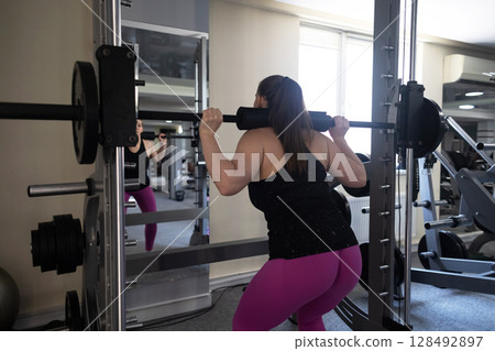 Plus size woman doing exercises on Smith Machine. Squats on a simulator with a barbell. Workout for weight loss 128492897