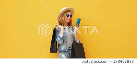 Beautiful happy young woman holding phone with black shopping bag, summer hat on yellow background 128492988