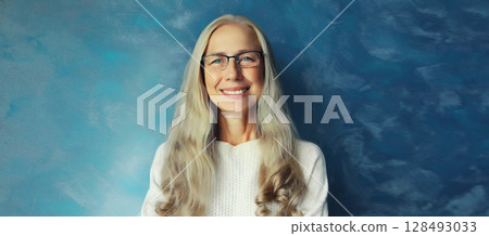 Portrait of happy smiling confident mature woman with eyeglasses looks at camera on blue background Portrait of happy smiling confident mature woman with eyeglasses looks at camera on blue background 128493033