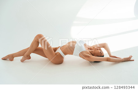Straightens the back up, sitting on the floor. Young woman with slim body type is in fitness clothes in the studio Straightens the back up, sitting on the floor. Young woman with slim body type is in fitness clothes in the studio 128493344