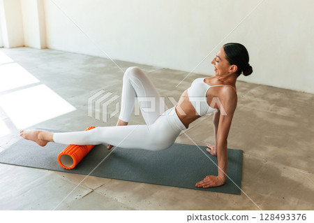 Orange colored foam roller. Young woman with slim body type is in fitness clothes in the studio Orange colored foam roller. Young woman with slim body type is in fitness clothes in the studio 128493376