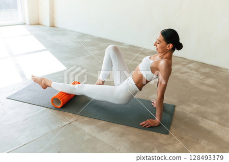 Using foam roller for exercises. Young woman with slim body type is in fitness clothes in the studio 128493379