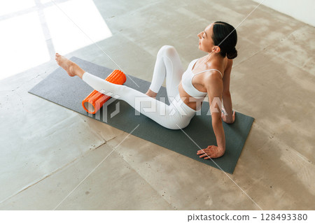 With orange colored foam roller. Young woman with slim body type is in fitness clothes in the studio 128493380