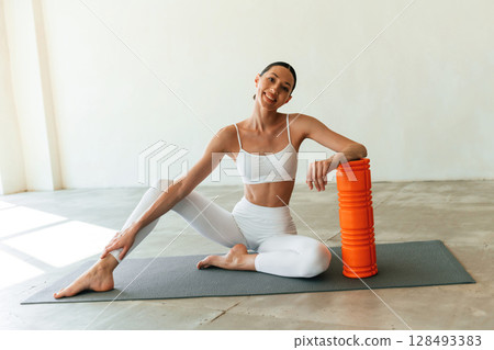 With orange colored foam roller. Young woman with slim body type is in fitness clothes in the studio 128493383
