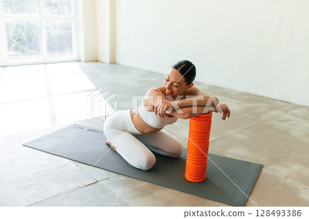 Sitting with foam roller. Young woman with slim body type is in fitness clothes in the studio 128493386