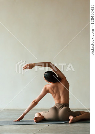 Flexible body, sitting, rear view. Young woman with slim body type is in fitness clothes in the studio Flexible body, sitting, rear view. Young woman with slim body type is in fitness clothes in the studio 128493433