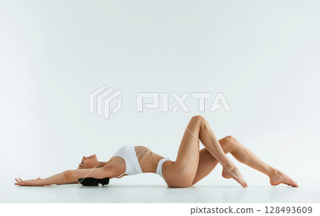 Side view, lying down. Young woman with slim body type is in fitness clothes in the studio Side view, lying down. Young woman with slim body type is in fitness clothes in the studio 128493609