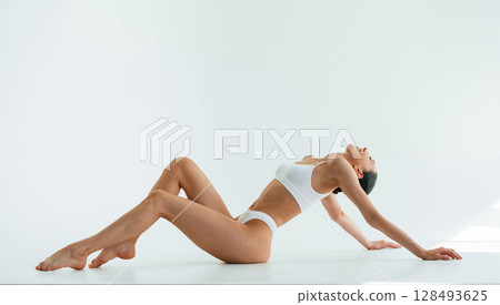 View from the side, on the floor. Young woman with slim body type is in fitness clothes in the studio View from the side, on the floor. Young woman with slim body type is in fitness clothes in the studio 128493625