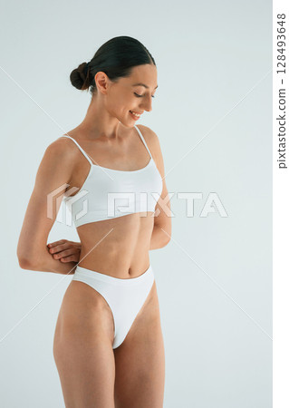 Hands behind the waist. Young woman with slim body type is in fitness clothes in the studio 128493648
