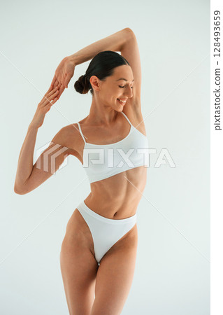 Relaxed, standing against white background. Young woman with slim body type is in fitness clothes in the studio Relaxed, standing against white background. Young woman with slim body type is in fitness clothes in the studio 128493659