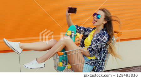 Stylish young woman taking selfie with phone, cool teenage girl in shopping trolley cart in the city Stylish young woman taking selfie with phone, cool teenage girl in shopping trolley cart in the city 128493830