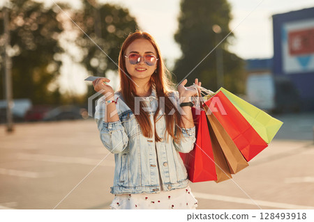 Discounts, with credit card in hands. Beautiful woman in casual clothes is holding shopping bags, outdoors Discounts, with credit card in hands. Beautiful woman in casual clothes is holding shopping bags, outdoors 128493918