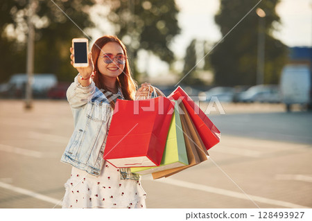Showing smartphone that is in the hand. Beautiful woman in casual clothes is holding shopping bags, outdoors 128493927