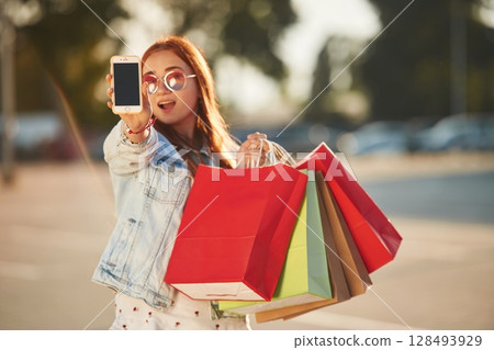 Showing smartphone that is in the hand. Beautiful woman in casual clothes is holding shopping bags, outdoors Showing smartphone that is in the hand. Beautiful woman in casual clothes is holding shopping bags, outdoors 128493929