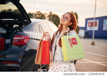 Standing near the car that is with the trunk open. Beautiful woman in casual clothes is holding shopping bags, outdoors Standing near the car that is with the trunk open. Beautiful woman in casual clothes is holding shopping bags, outdoors 128493938