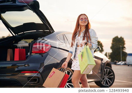 Against the car with trunk opened. Beautiful woman in casual clothes is holding shopping bags, outdoors Against the car with trunk opened. Beautiful woman in casual clothes is holding shopping bags, outdoors 128493940