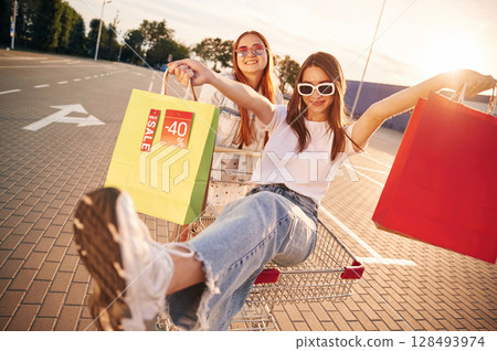 Against sunlight, riding in the shop cart. Two beautiful women in casual clothes are holding shopping bags, outdoors 128493974