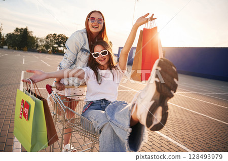 Friends are enjoying riding on shop cart. Two beautiful women in casual clothes are holding bags, outdoors Friends are enjoying riding on shop cart. Two beautiful women in casual clothes are holding bags, outdoors 128493979