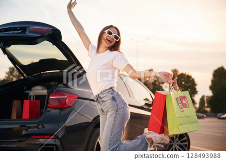 Against the car with trunk opened. Beautiful woman in casual clothes is holding shopping bags, outdoors Against the car with trunk opened. Beautiful woman in casual clothes is holding shopping bags, outdoors 128493988