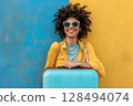 Smiling woman wearing sunglasses with blue suitcase standing against blue and yellow wall 128494074