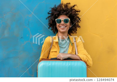Smiling woman wearing sunglasses with blue suitcase standing against blue and yellow wall 128494074