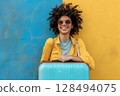 Smiling woman wearing sunglasses with blue suitcase standing against blue and yellow wall 128494075