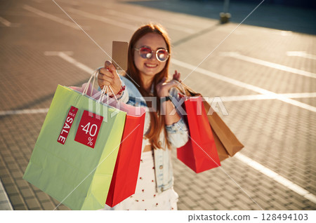 Discounts, with credit card in hands. Beautiful woman in casual clothes is holding shopping bags, outdoors 128494103