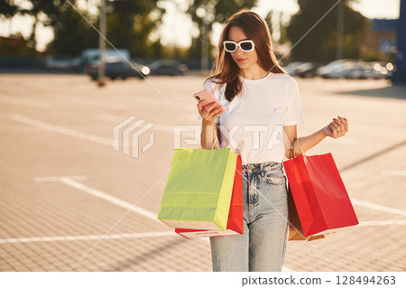 Using smartphone. Beautiful woman in casual clothes is holding shopping bags, outdoors Using smartphone. Beautiful woman in casual clothes is holding shopping bags, outdoors 128494263