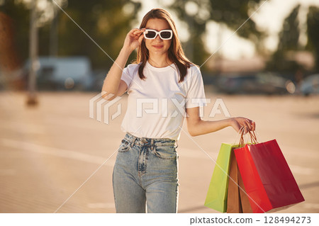 Wearing the sunglasses, standing. Beautiful woman in casual clothes is holding shopping bags, outdoors 128494273