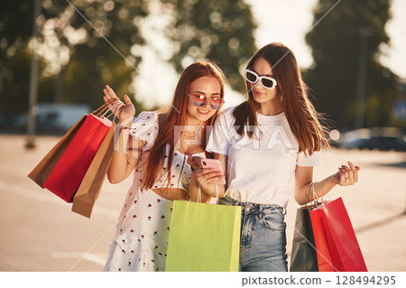 Two beautiful women in casual clothes are holding shopping bags, outdoors Two beautiful women in casual clothes are holding shopping bags, outdoors 128494295
