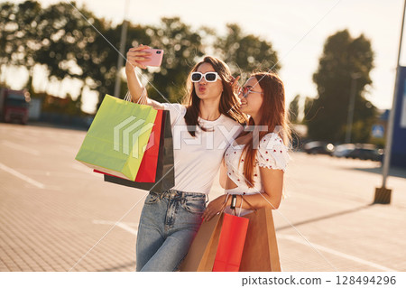 Two beautiful women in casual clothes are holding shopping bags, outdoors 128494296