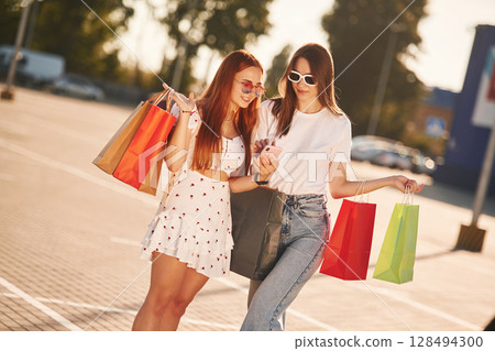 Excited by content in the smartphone. Two beautiful women in casual clothes are holding shopping bags, outdoors 128494300