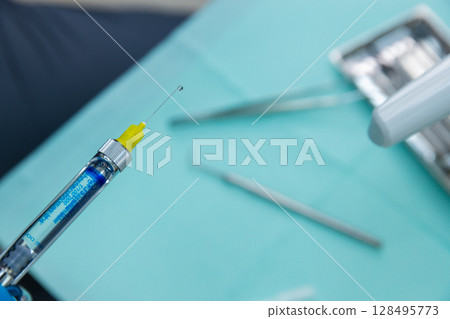 Close-Up of a Syringe with a Yellow Cap and Blue Liquid Ready for Injection on a Medical Table Surrounded by Surgical Instruments Providing Clinical Context for Healthcare Settings 128495773