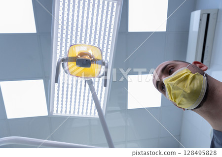 A Focused Dental Professional in a Clinical Environment Preparing for Treatment Under Bright Overhead Lights and Precautionary Gear 128495828